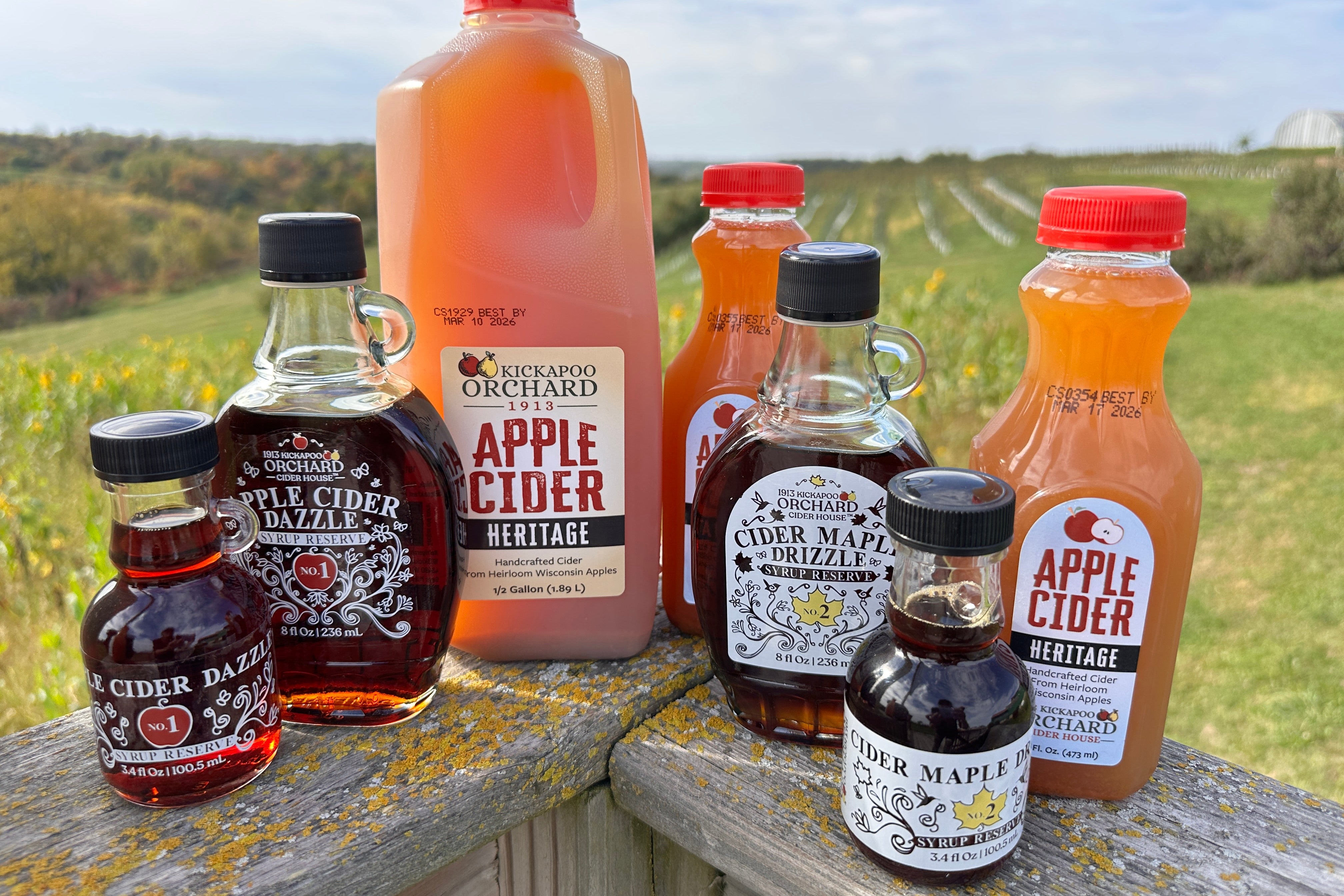 Collection of apple cider and maple syrup bottles on a wooden surface with a scenic background.